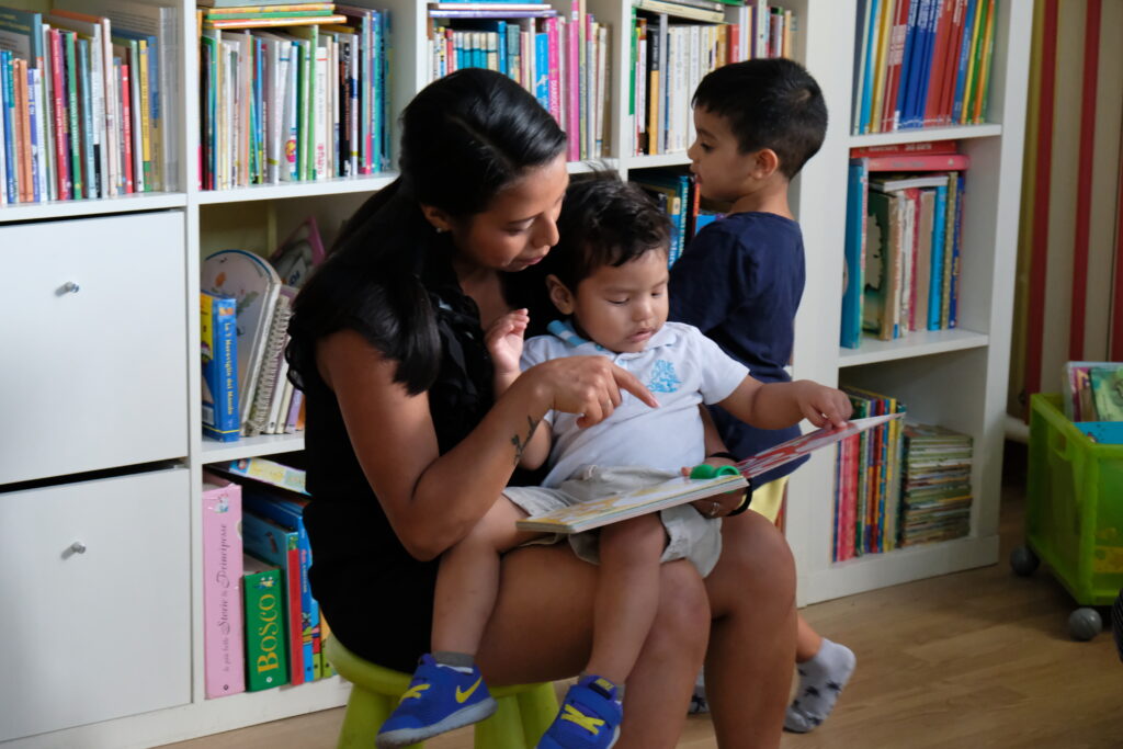 Una mamma e un bambino leggono un libro, un altro bambino è in piedi davanti a una libreria piena di libri.